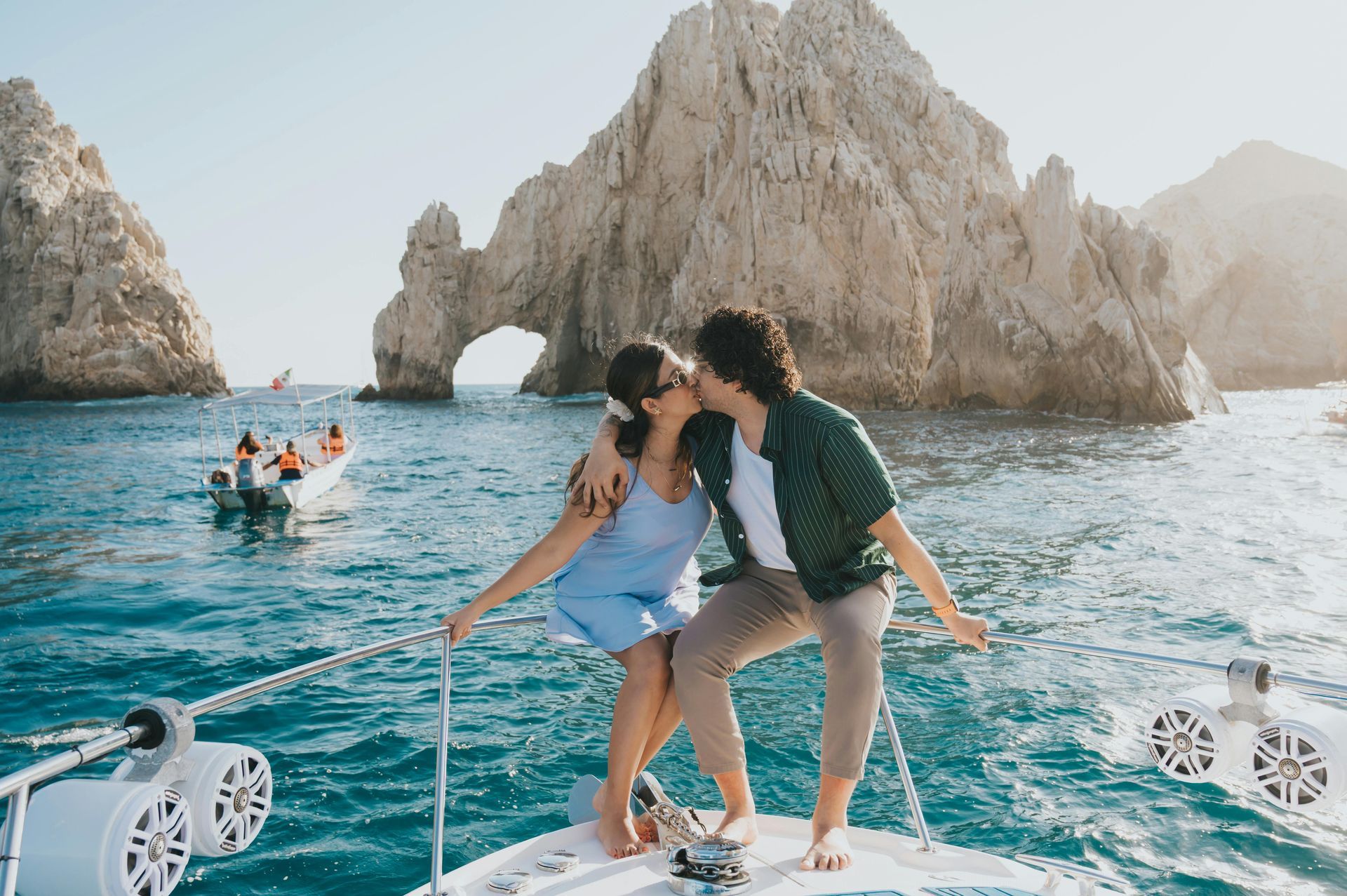 A couple kisses on the bow of a boat in front of the iconic rock arch in Cabo San Lucas, with a small boat nearby.