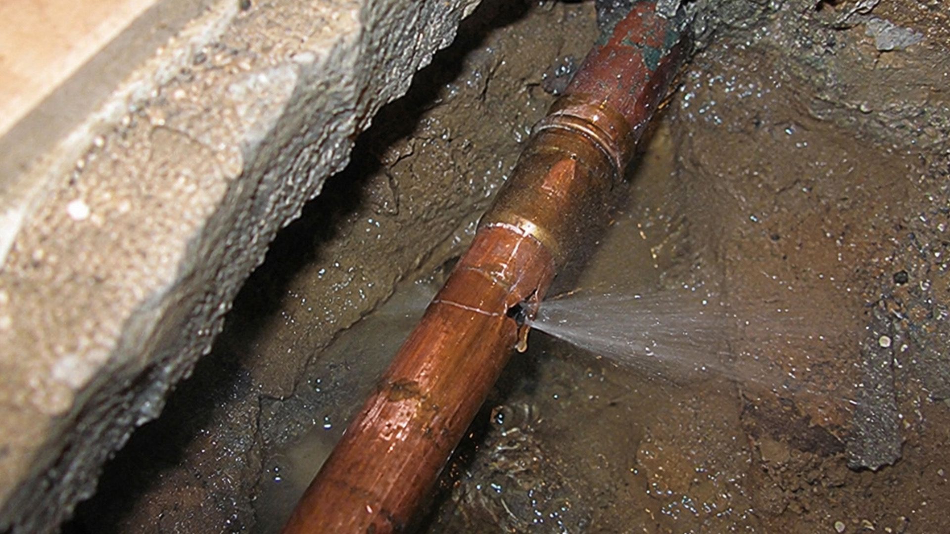 Leaking copper pipe inside a wall cavity with water spraying onto wet dirt and concrete.