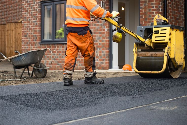 Construction worker in orange safety gear using a yellow roller to compact asphalt driveway in front of a brick house.