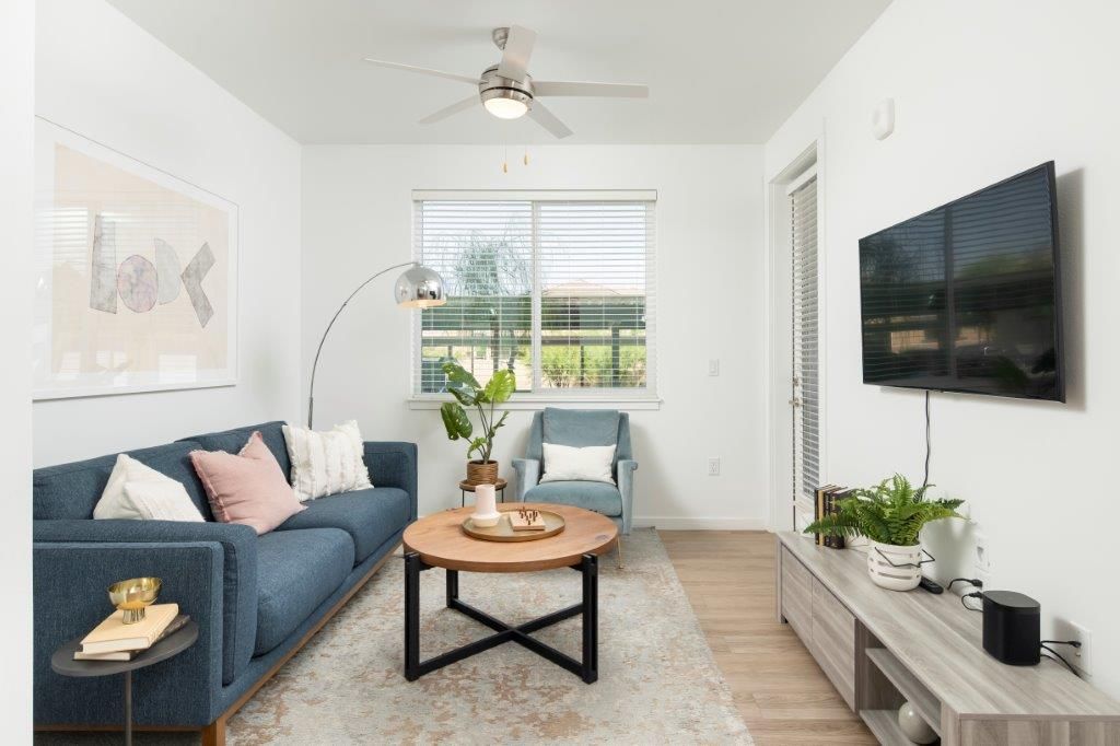 Bright modern apartment living room with blue sofa, round coffee table, wall TV, and potted plants.