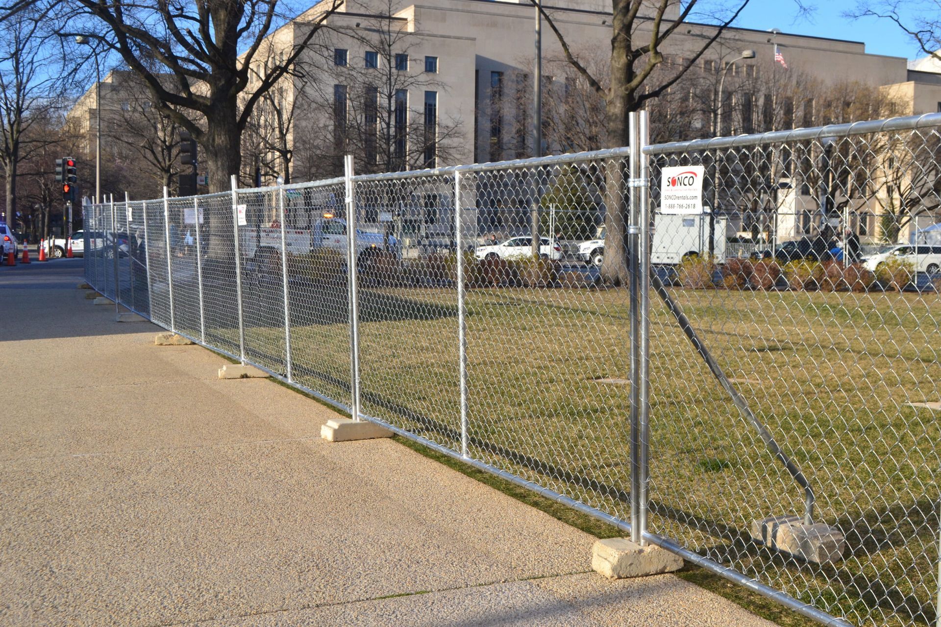 Chain-link fence on concrete, separating a grassy area from a sidewalk; a building is visible in the background.