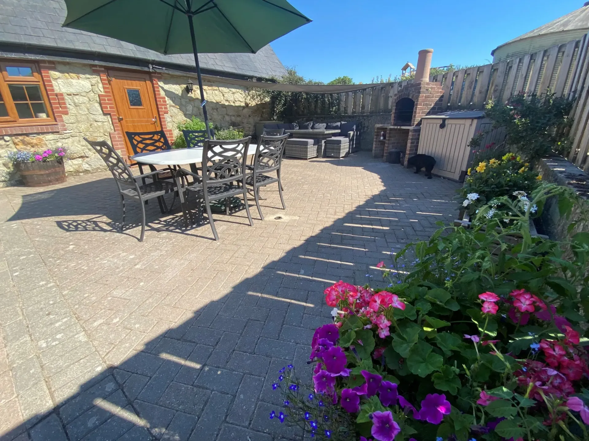 A stone patio featuring a dining set under an umbrella, an outdoor brick fireplace, and vibrant potted flowers.