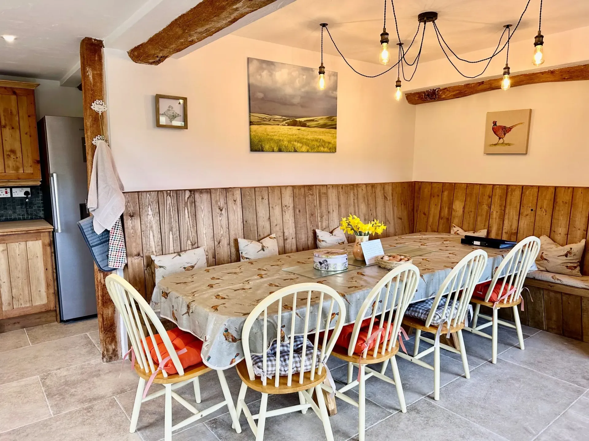 Dining area with a long table, white wooden chairs, and rustic wood-paneled booth seating against a white wall.