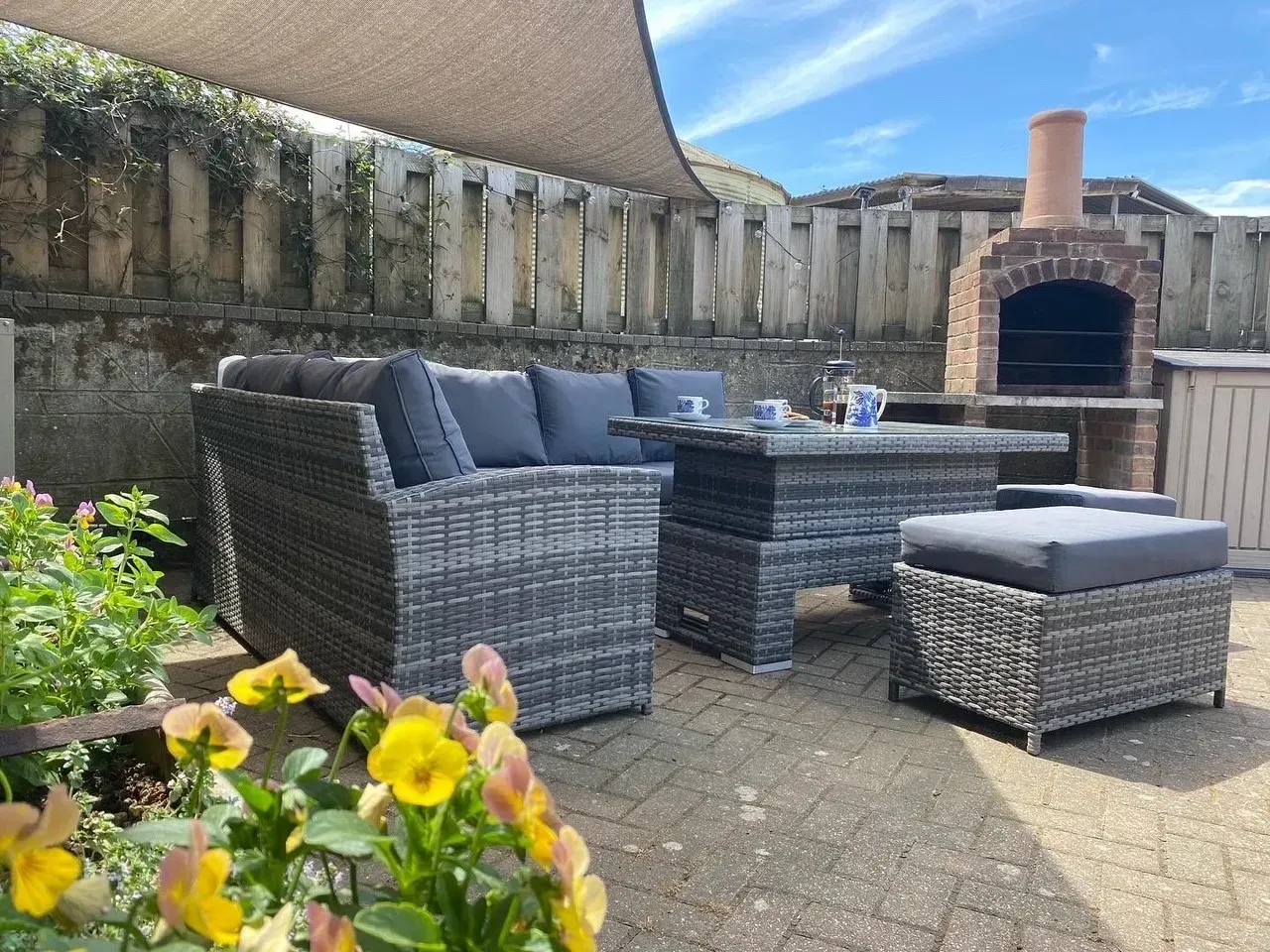 A gray wicker patio sofa set with dark cushions on a paved garden area near a brick outdoor fireplace and wooden fence.
