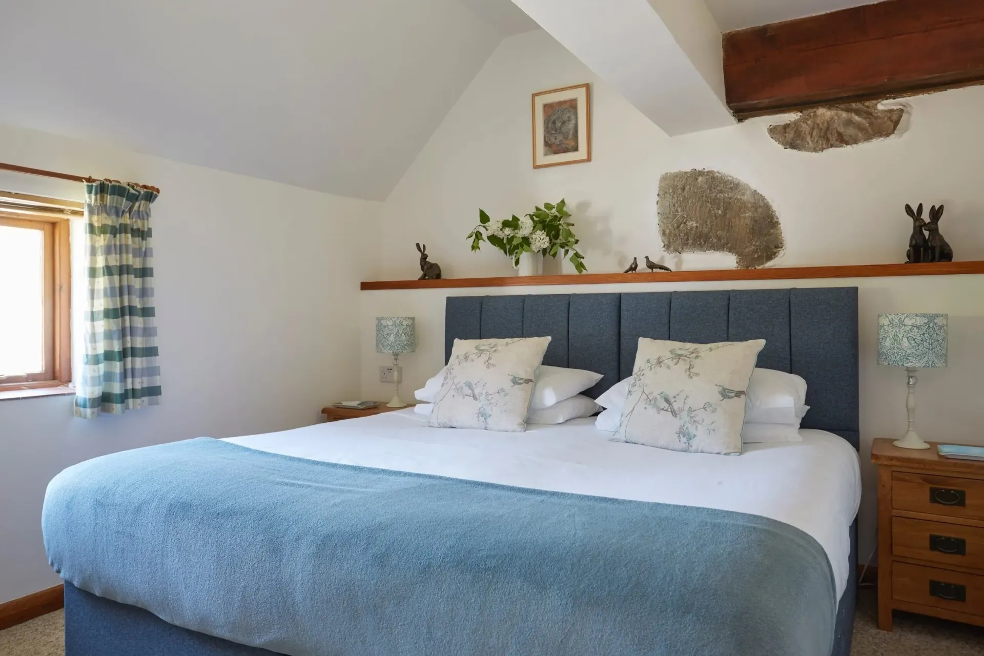 A bedroom with a bed featuring a blue headboard and throw, wooden nightstands, and a shelf with plants above the bed.