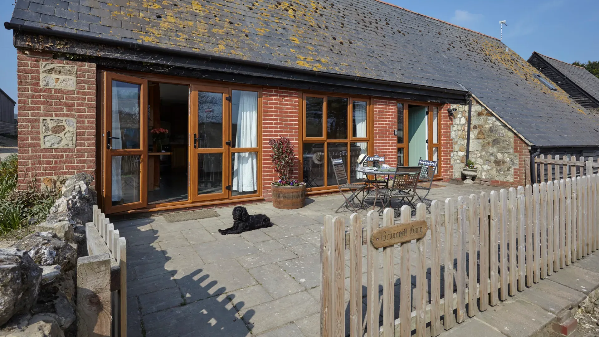 A brick cottage with large glass doors, a stone patio, a small fence, and a black dog lying in the sun.