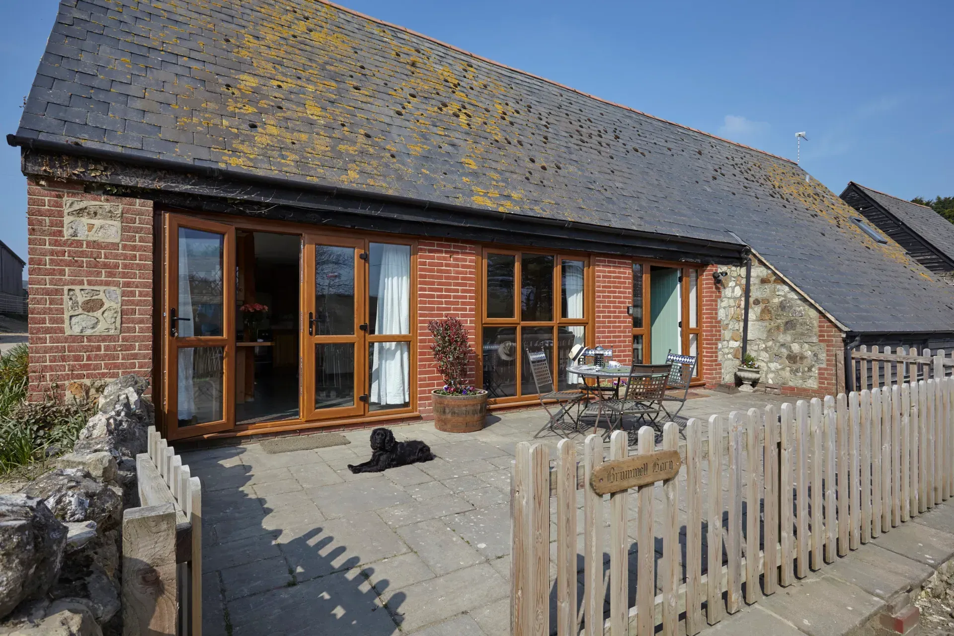 A brick holiday cottage with a slate roof, patio, and picket fence features large glass doors and a black dog resting.