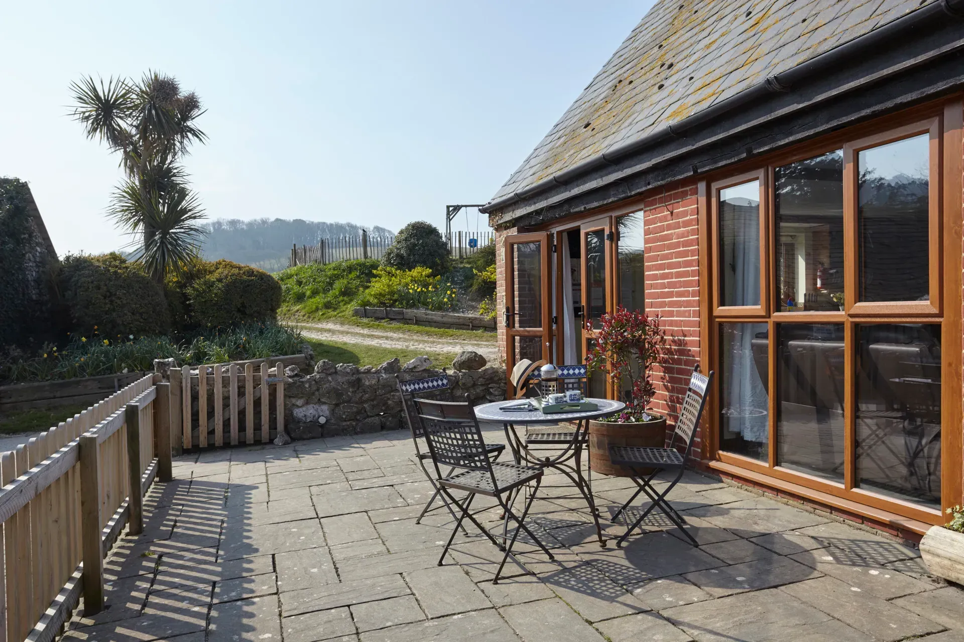 A paved patio with a bistro set outside a brick cottage, featuring wooden fencing and a countryside view in the distance.