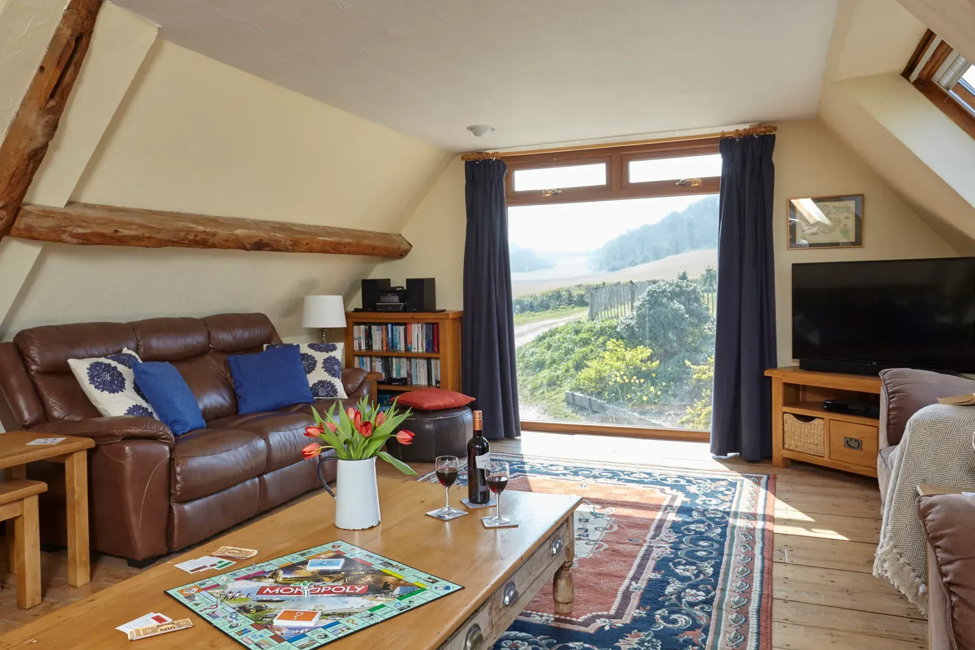A cozy attic living room with a brown leather sofa, wooden coffee table with a board game, and a large window with a view.