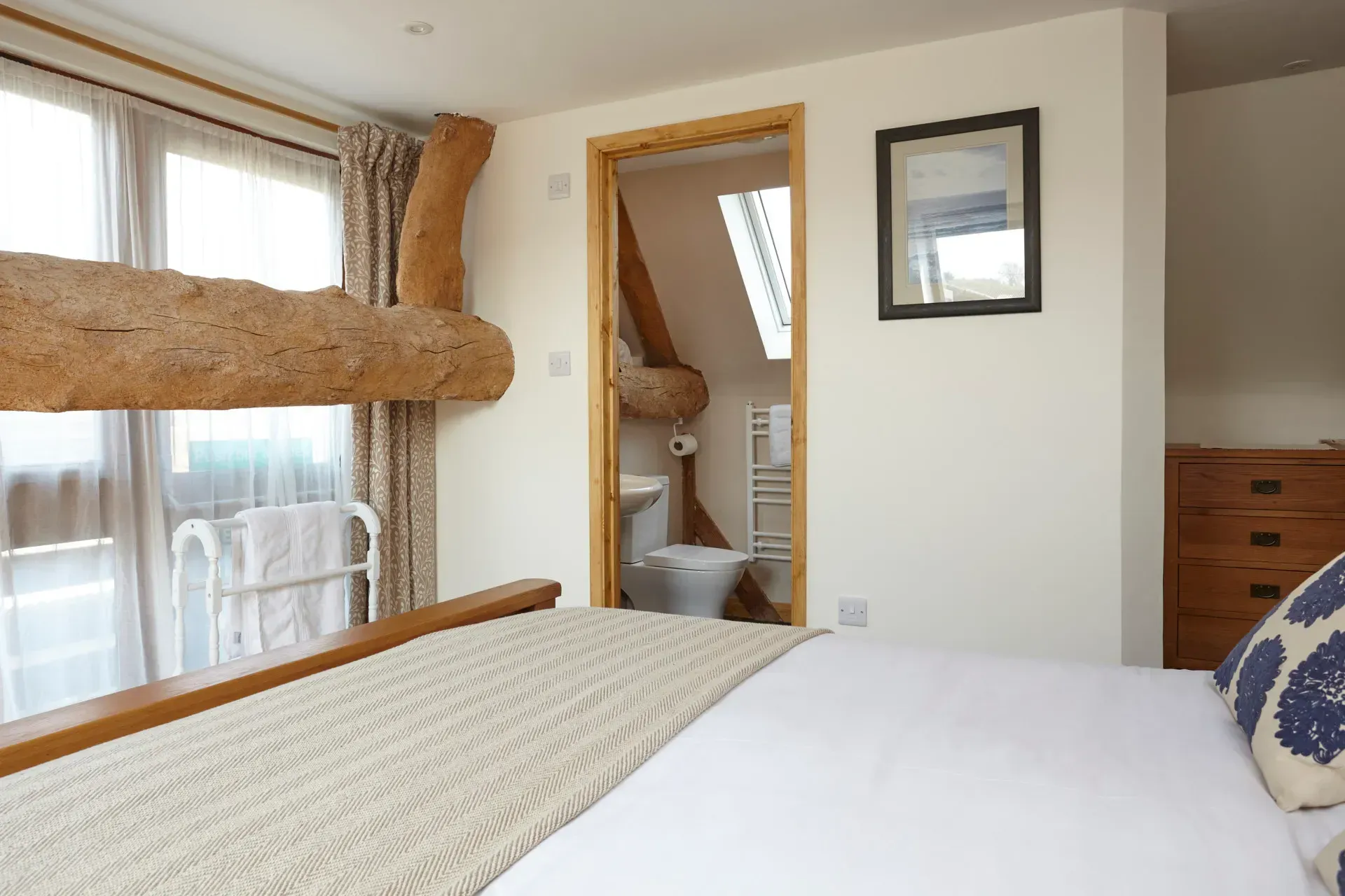 A bedroom with a wooden log beam feature, a white bed, and a doorway leading to a small en-suite bathroom with a skylight.