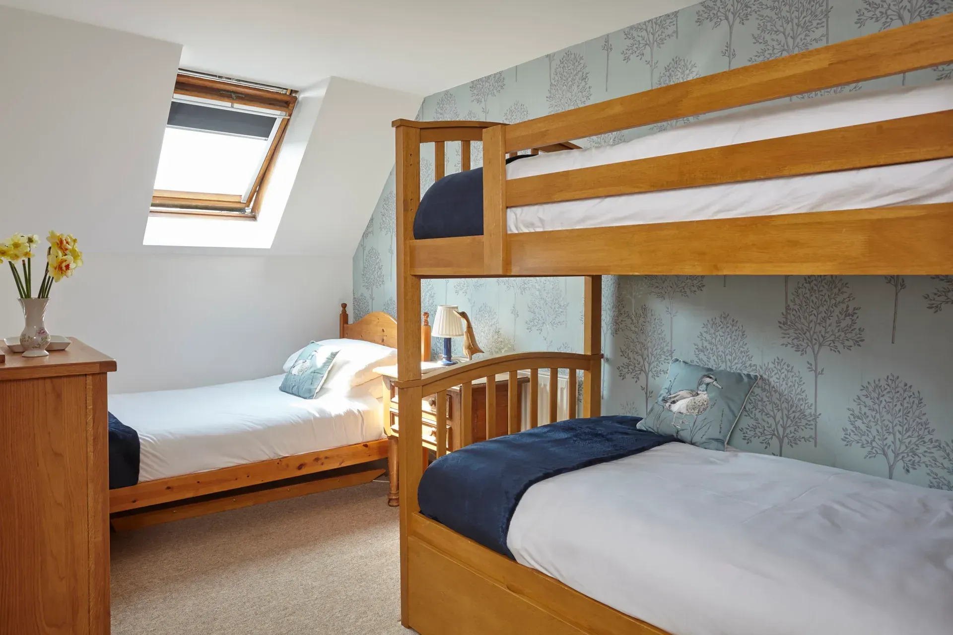 A bright bedroom with a bunk bed, a twin bed, wooden furniture, and light blue patterned wallpaper under a skylight.
