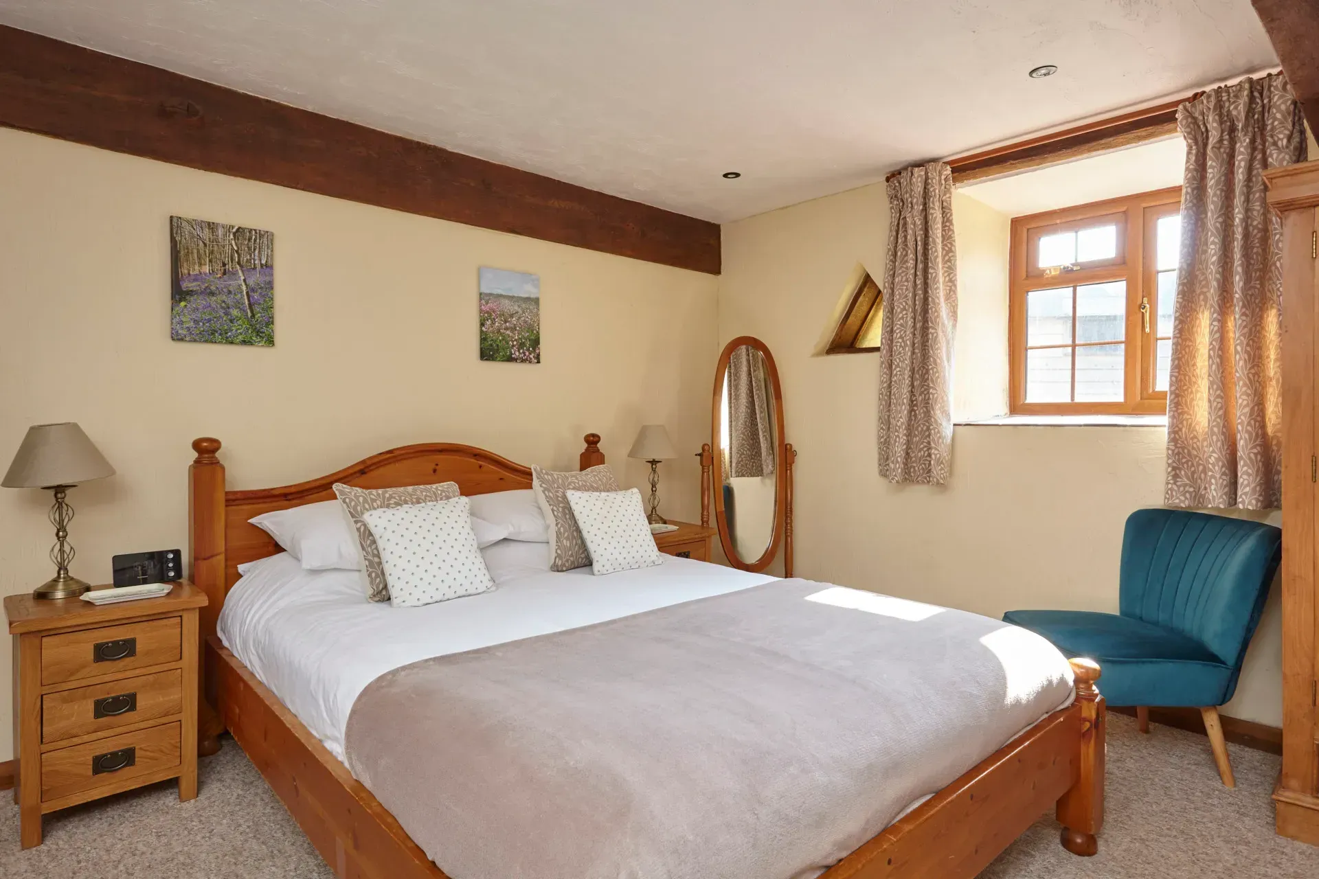 A bedroom with a wooden double bed, two bedside tables with lamps, a standing mirror, and a blue armchair by the window.