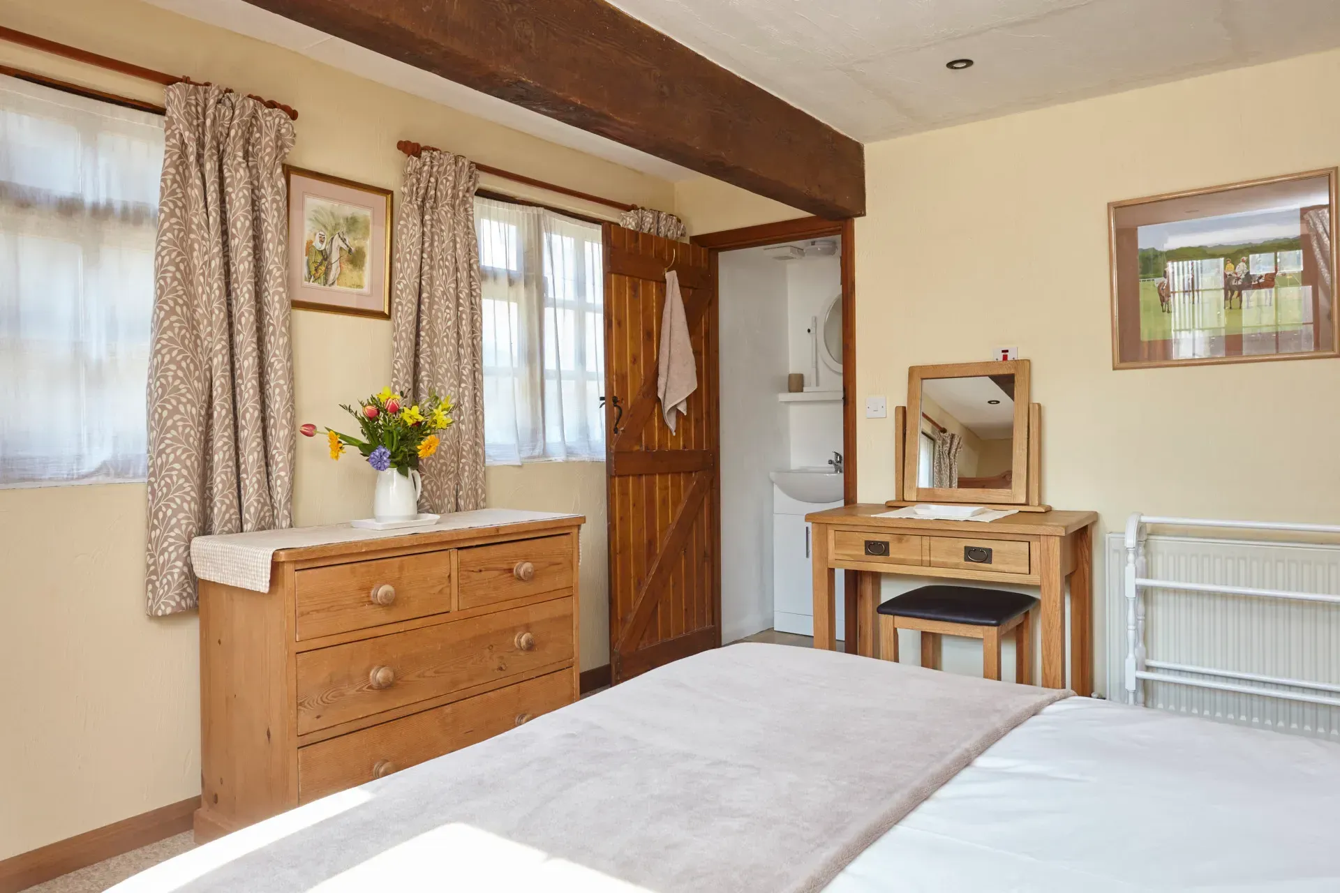 A bedroom with a wooden dresser, a vanity table with a mirror, a bed, and a door leading to a bathroom.
