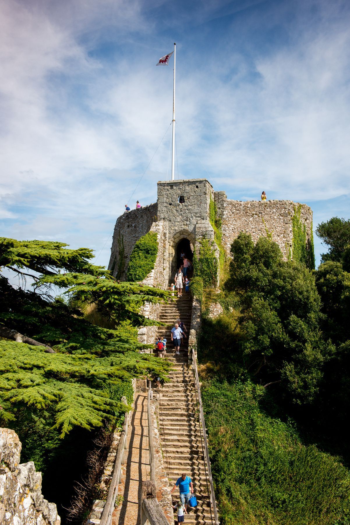 A stone castle tower atop a grassy hill, reached by a long, steep staircase with visitors climbing toward the entrance.