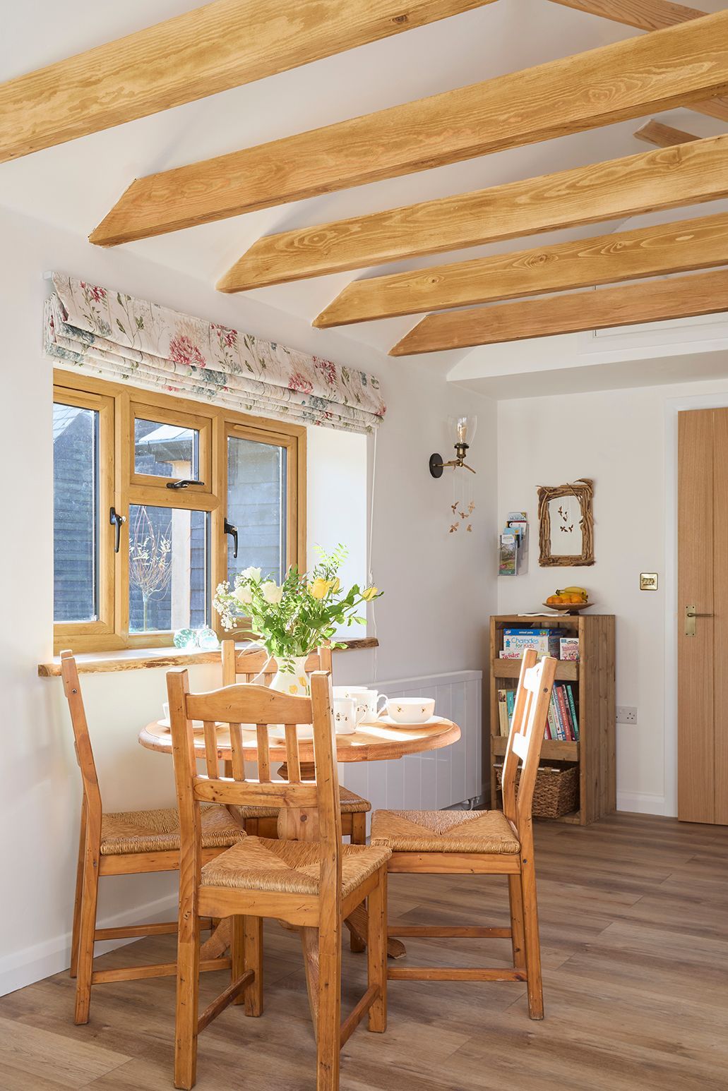 A small wooden dining set sits in a sunlit room with exposed ceiling beams, a window with floral curtains, and a shelf.