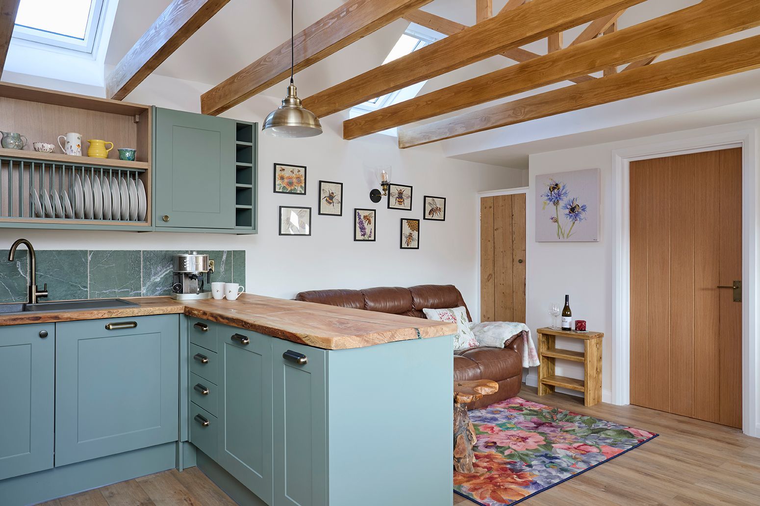 A cozy kitchen and living area featuring sage green cabinets, a wood countertop, exposed beams, and a floral area rug.