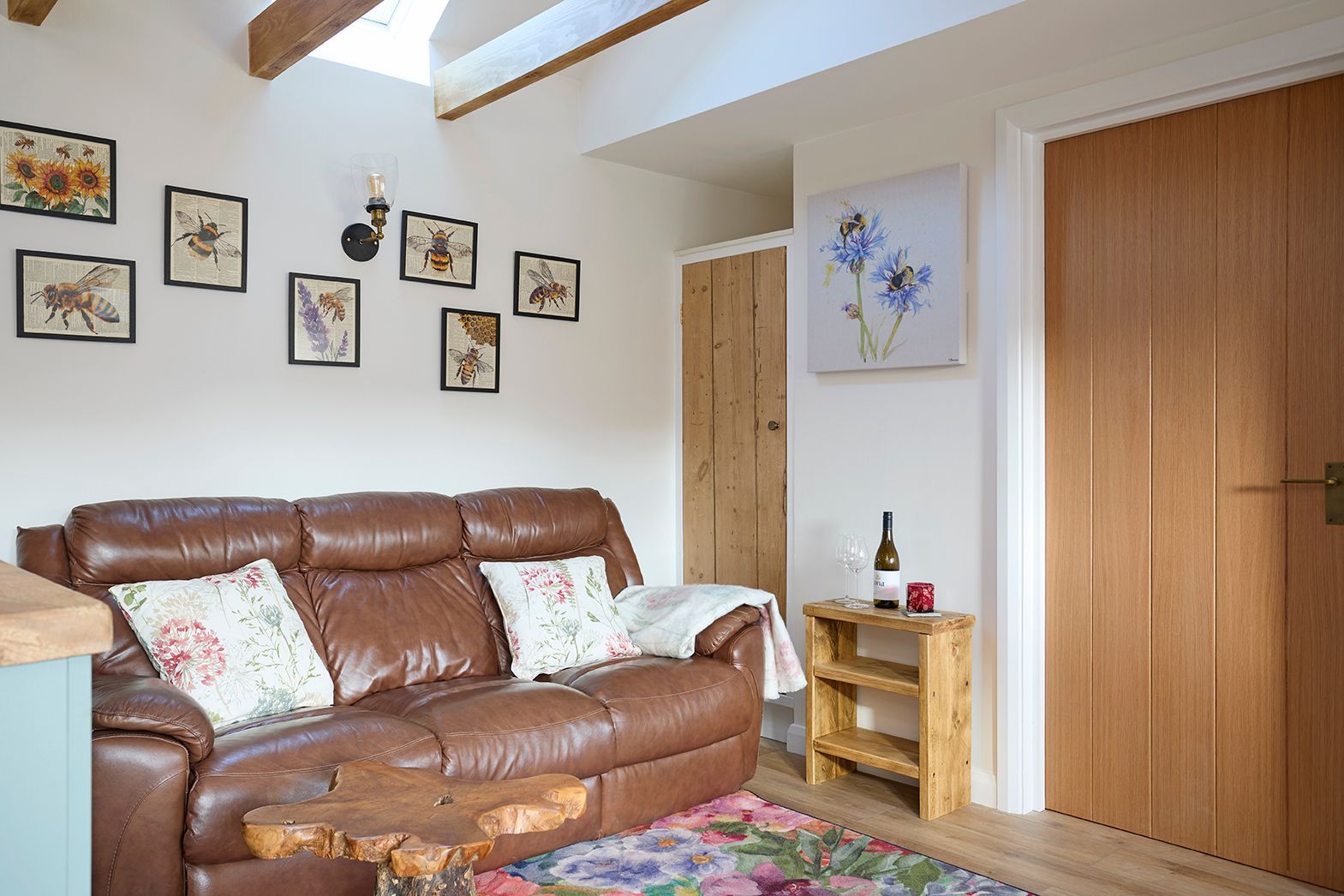 A brown leather sofa with floral pillows sits in a room with framed bee art, a wooden side table, and a wooden door.