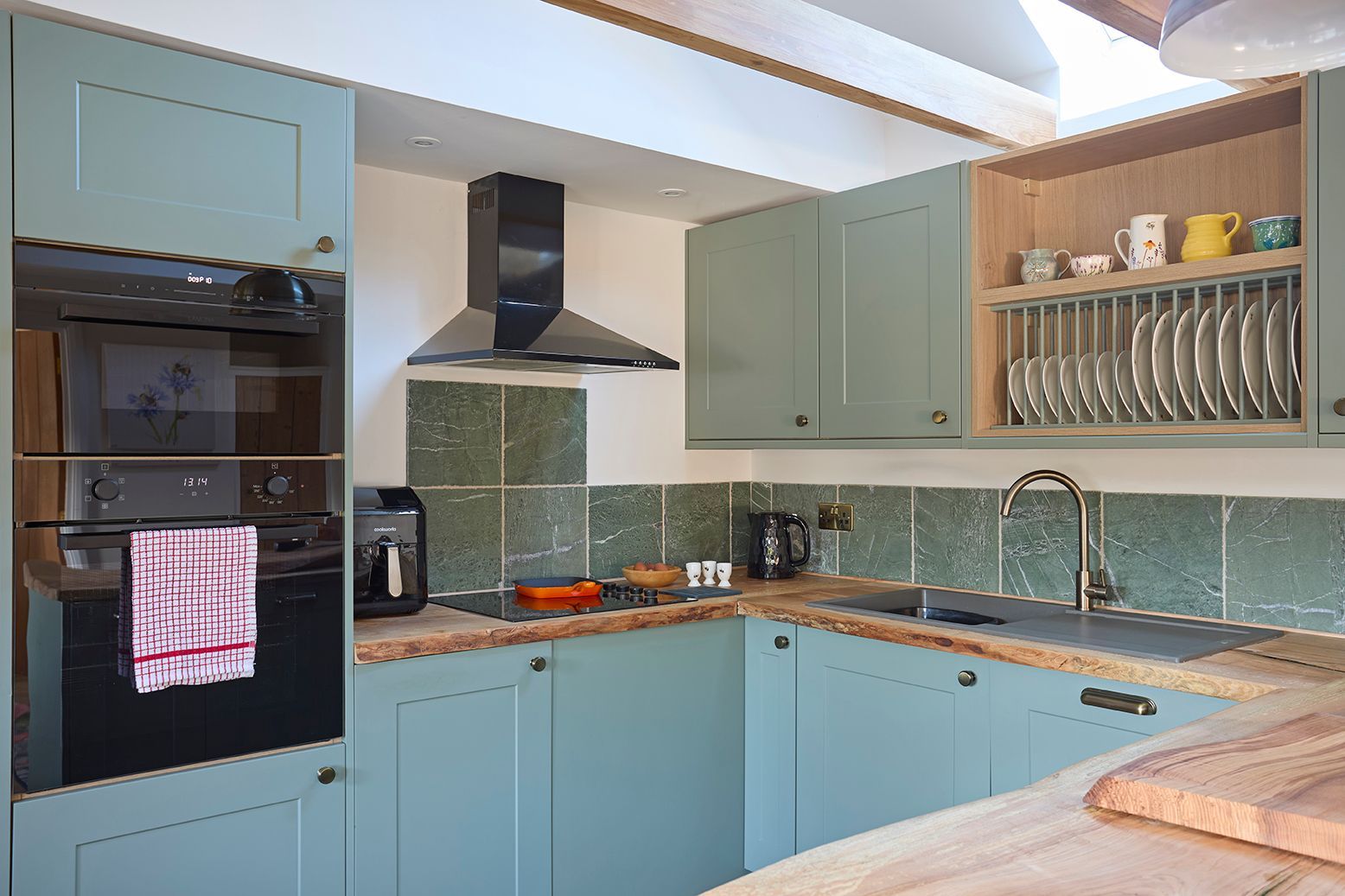 A modern kitchen with sage green cabinets, wood countertops, integrated appliances, and a slate-tiled backsplash.