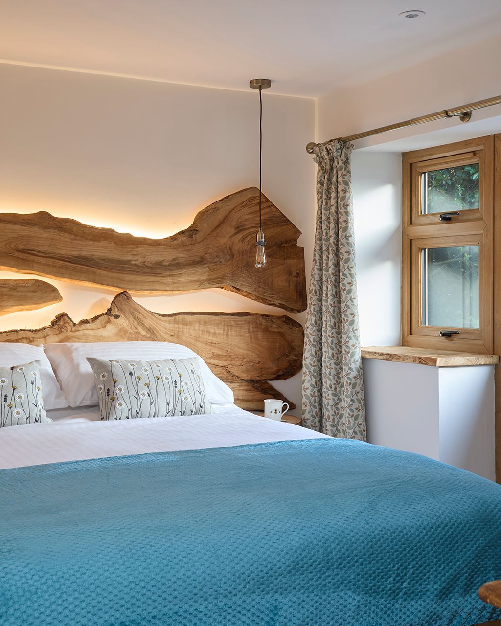 A bedroom with a live-edge wooden headboard illuminated by warm backlighting, a blue textured blanket, and a window.