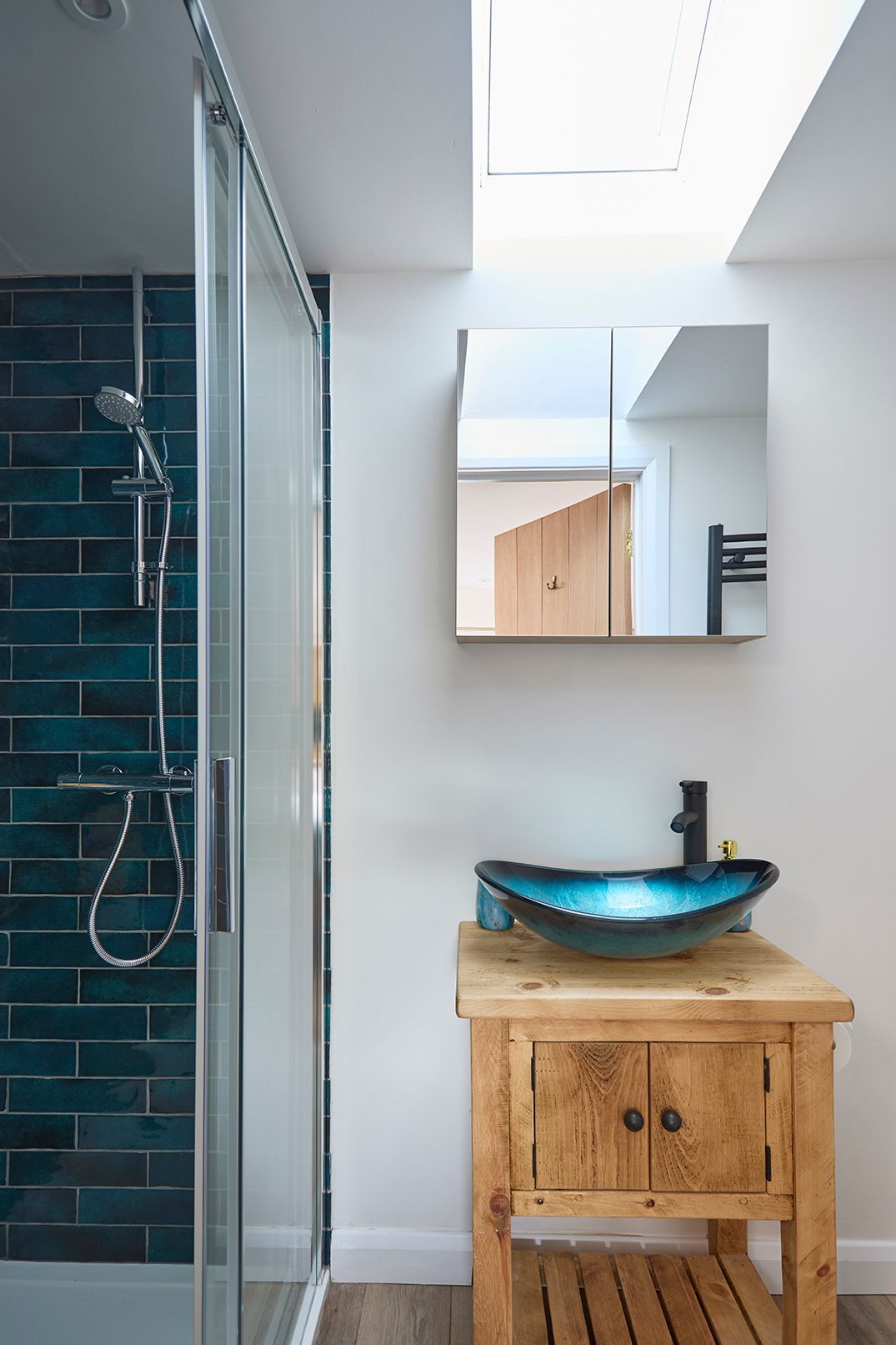 A bathroom with a rustic wooden vanity, a blue glass vessel sink, a walk-in shower with dark blue tiles, and a skylight.