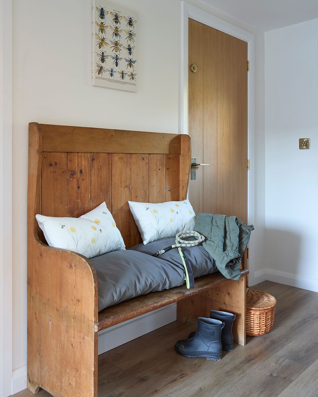 A wooden bench with gray cushions and patterned pillows sits in an entryway with boots nearby and art on the wall.