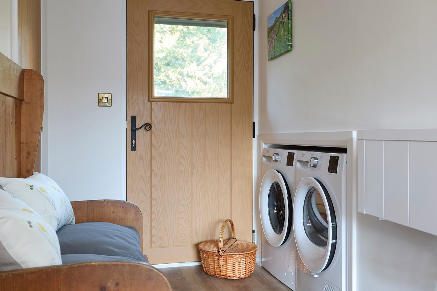 A utility room with two front-loading machines, a wicker laundry basket, a wooden door, and a couch in the foreground.