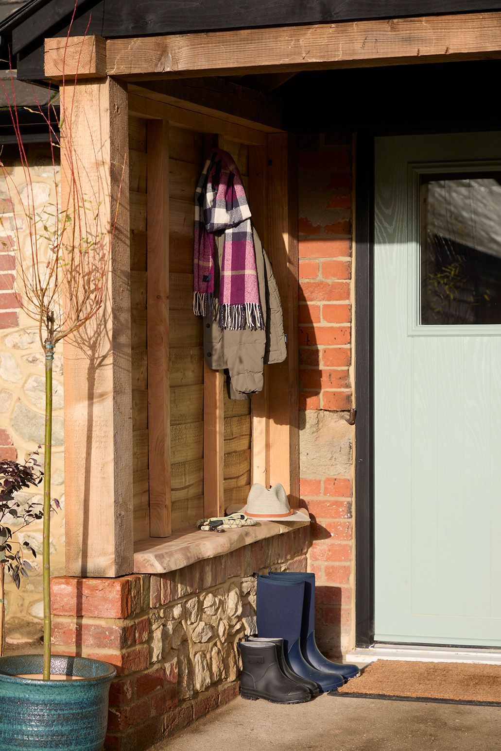 A small outdoor porch entrance featuring a pale green door, stone and brick wall, wooden post, hanging coats, and boots.