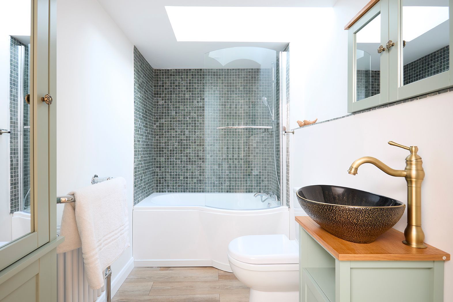 A modern bathroom with sage green cabinetry, a patterned vessel sink, gold faucet, and a shower with dark mosaic tiles.