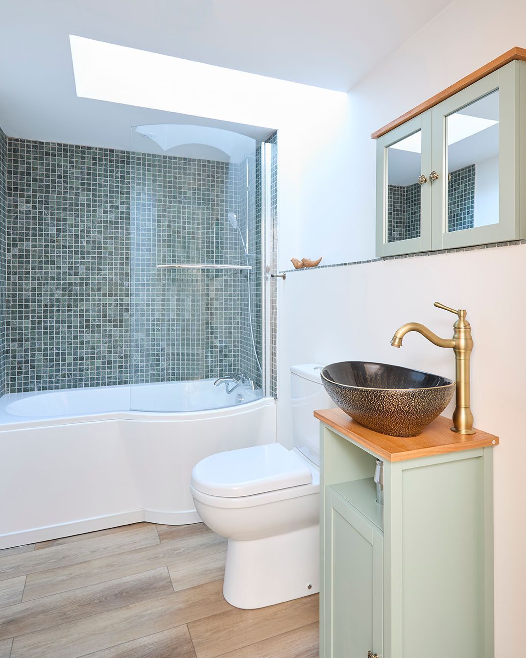 A bathroom with a tiled bathtub shower combo, a white toilet, and a light green vanity with a vessel sink and gold faucet.