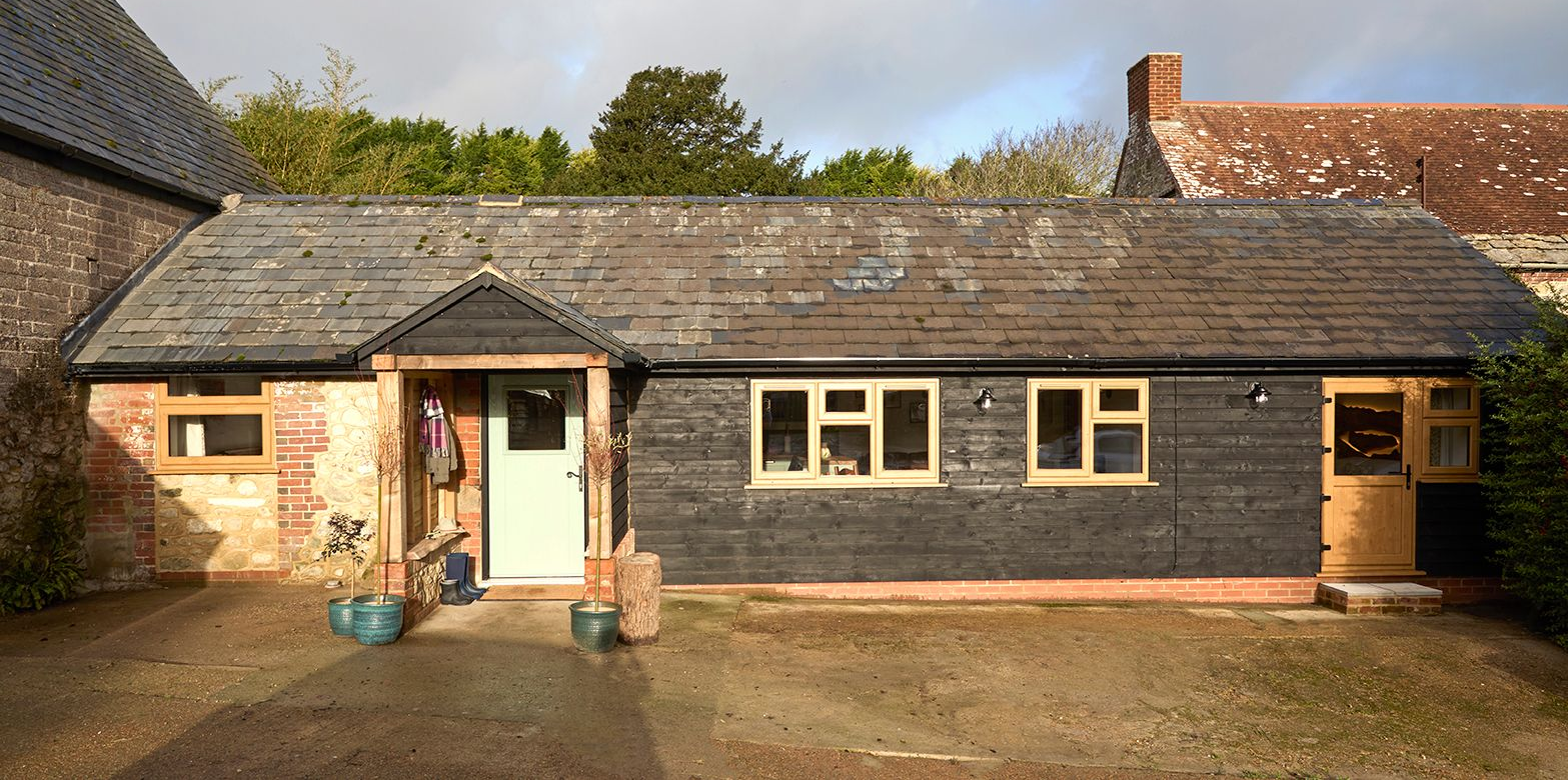 Single-story building with a light-colored stone section, dark wood siding, a slate roof, and wooden-framed windows.