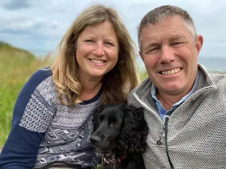 A woman and a man smile outdoors with a black spaniel between them, set against a backdrop of coastal greenery and sea.