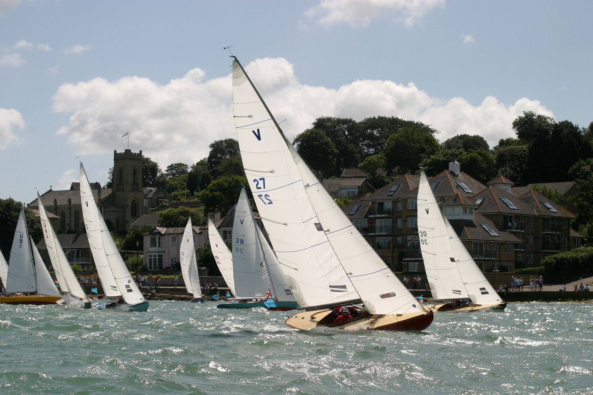 A regatta of sailing boats with white sails moves across a sparkling body of water toward a shoreline with buildings.