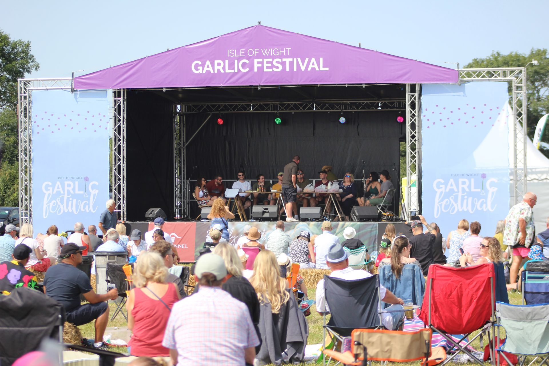 People sit in lawn chairs watching a panel discussion on a stage at the Isle of Wight Garlic Festival under a purple sign.