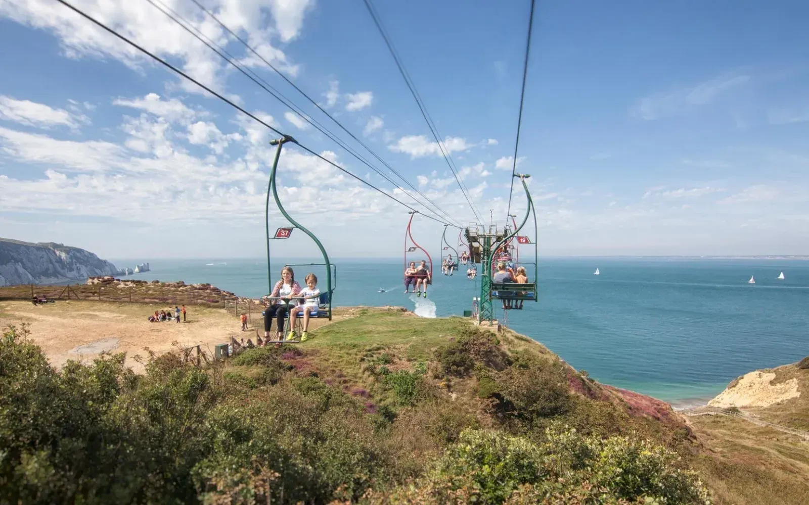 A chairlift carries passengers over a grassy cliff overlooking a blue sea under a bright, partly cloudy sky.