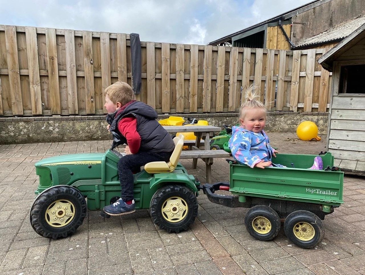 Two children ride toy vehicles in a backyard, one driving a green tractor and one in a green trailer.