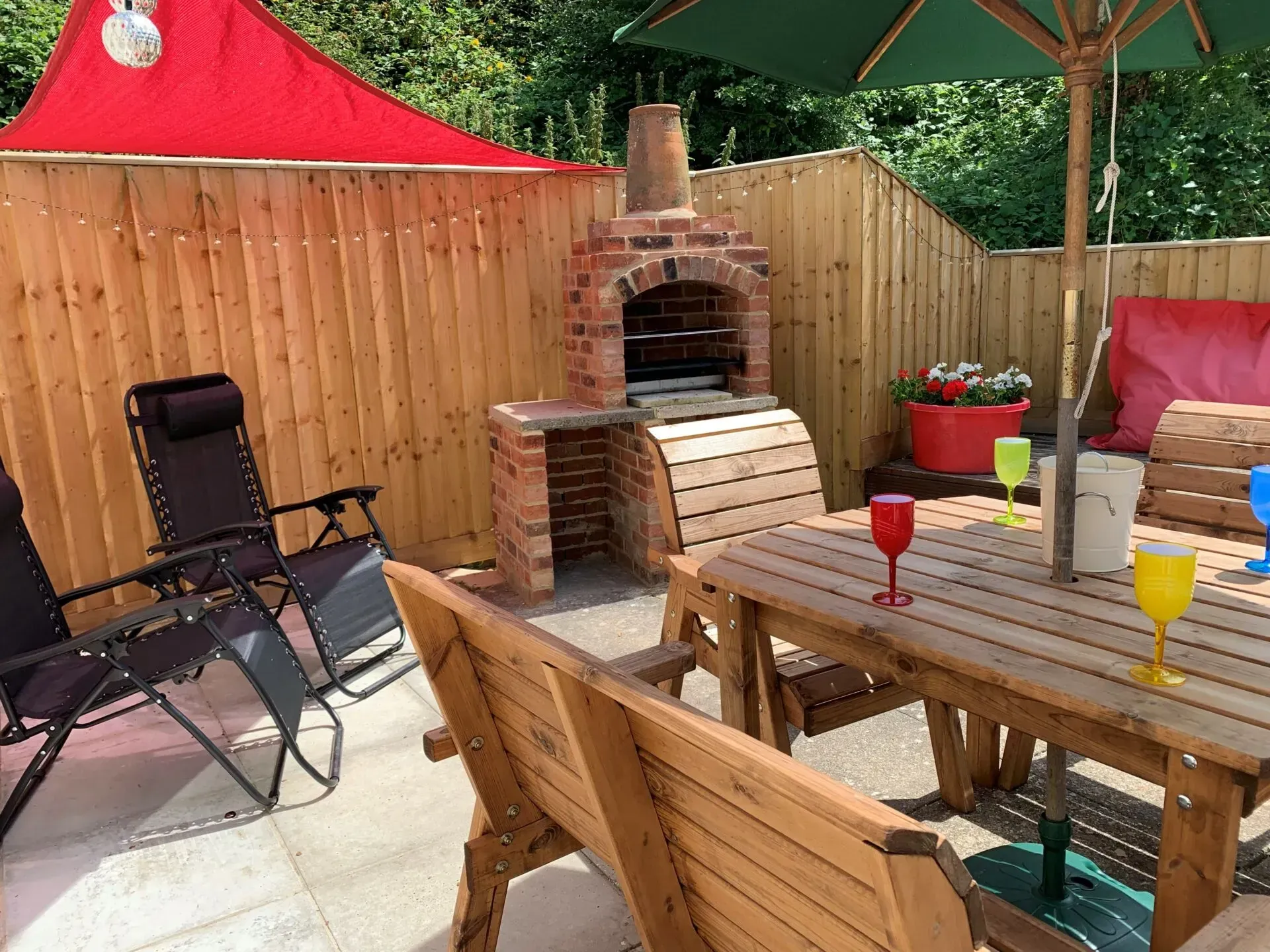 A patio area with a wooden table, chairs, a brick barbecue, and a red shade sail against wooden fence panels.