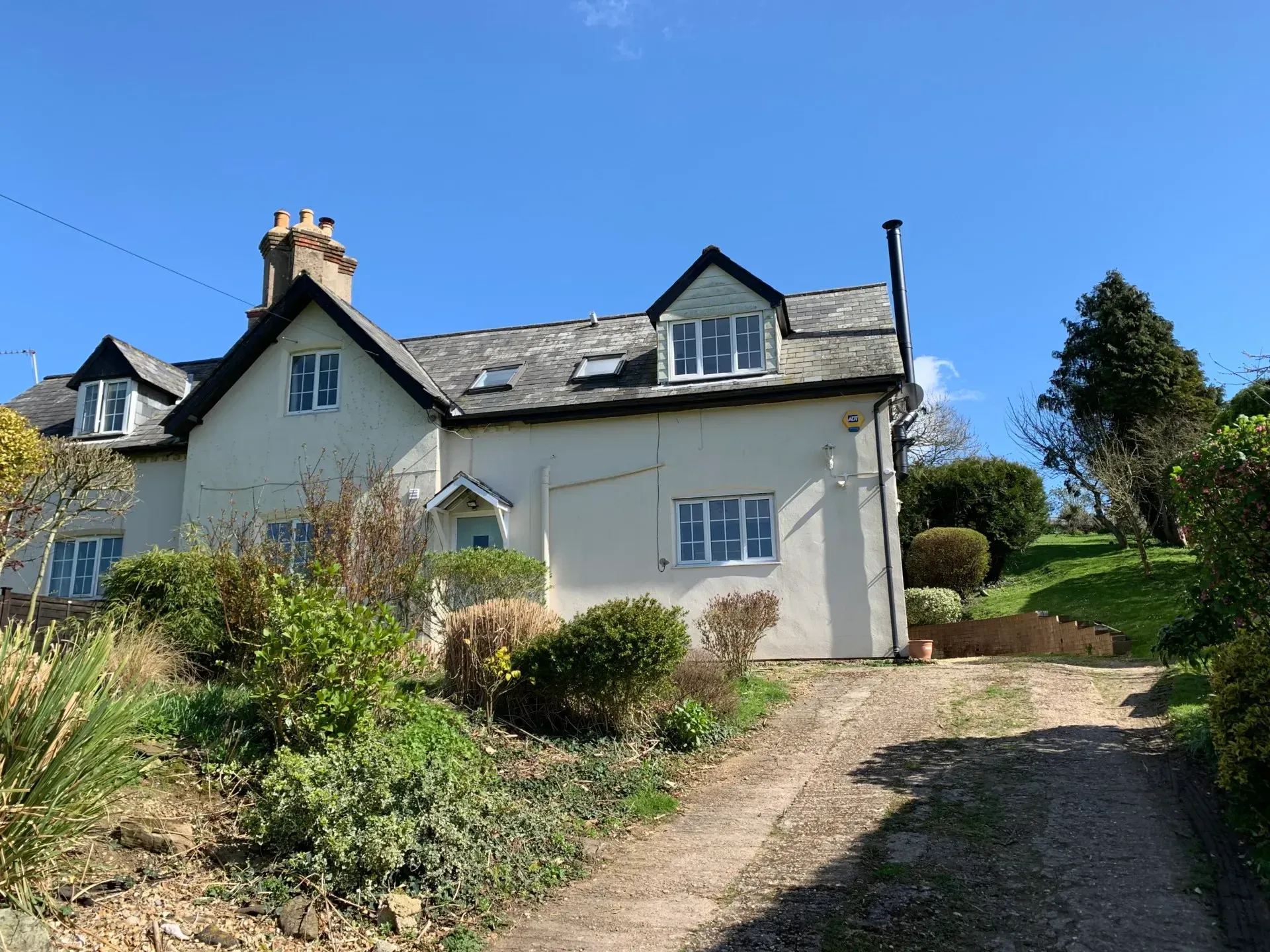 A white two-story cottage with a gray roof and dormer windows, surrounded by a garden and a gravel driveway.