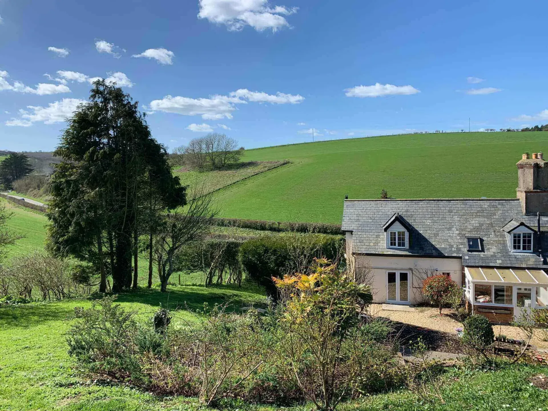 A stone cottage with a slate roof set against a rolling green hill under a bright blue sky with scattered clouds.