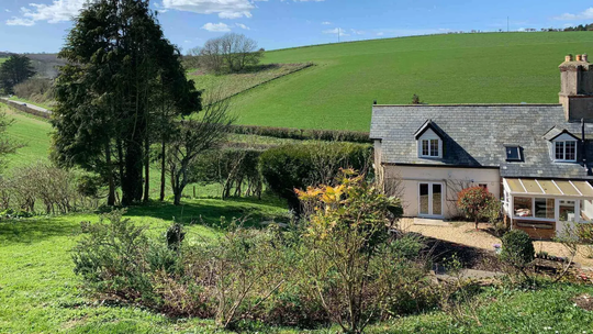 A stone cottage with a slate roof sits nestled in a lush, green, rolling hillside landscape under a bright blue sky.
