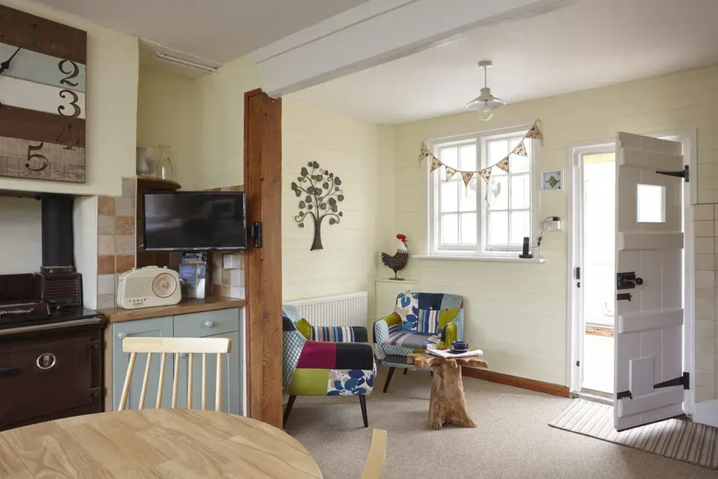 A cozy, sunlit room with two patterned armchairs, a wooden side table, a round table, and an open door leading outside.