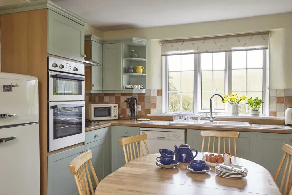 A bright, cozy kitchen with sage cabinets, a white retro fridge, a wooden table with tea service, and a large window.