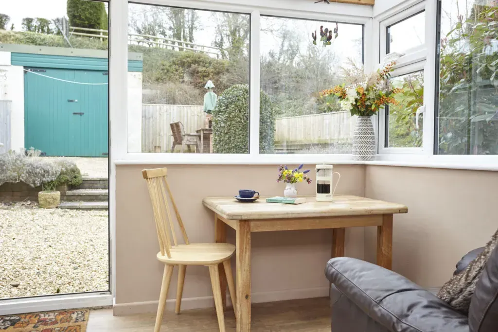 A sunroom with a wooden table, a light wood spindle chair, a vase of flowers, and a dark leather sofa.