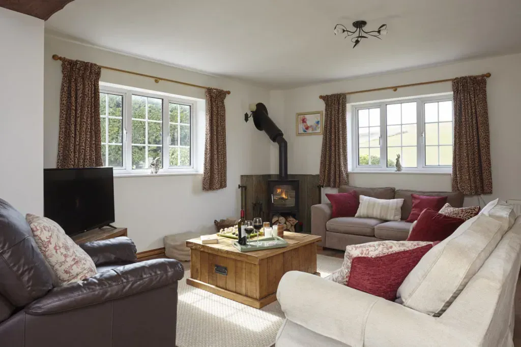 A cozy living room with a wood-burning stove, white sofa, armchairs, coffee table, and windows with patterned curtains.
