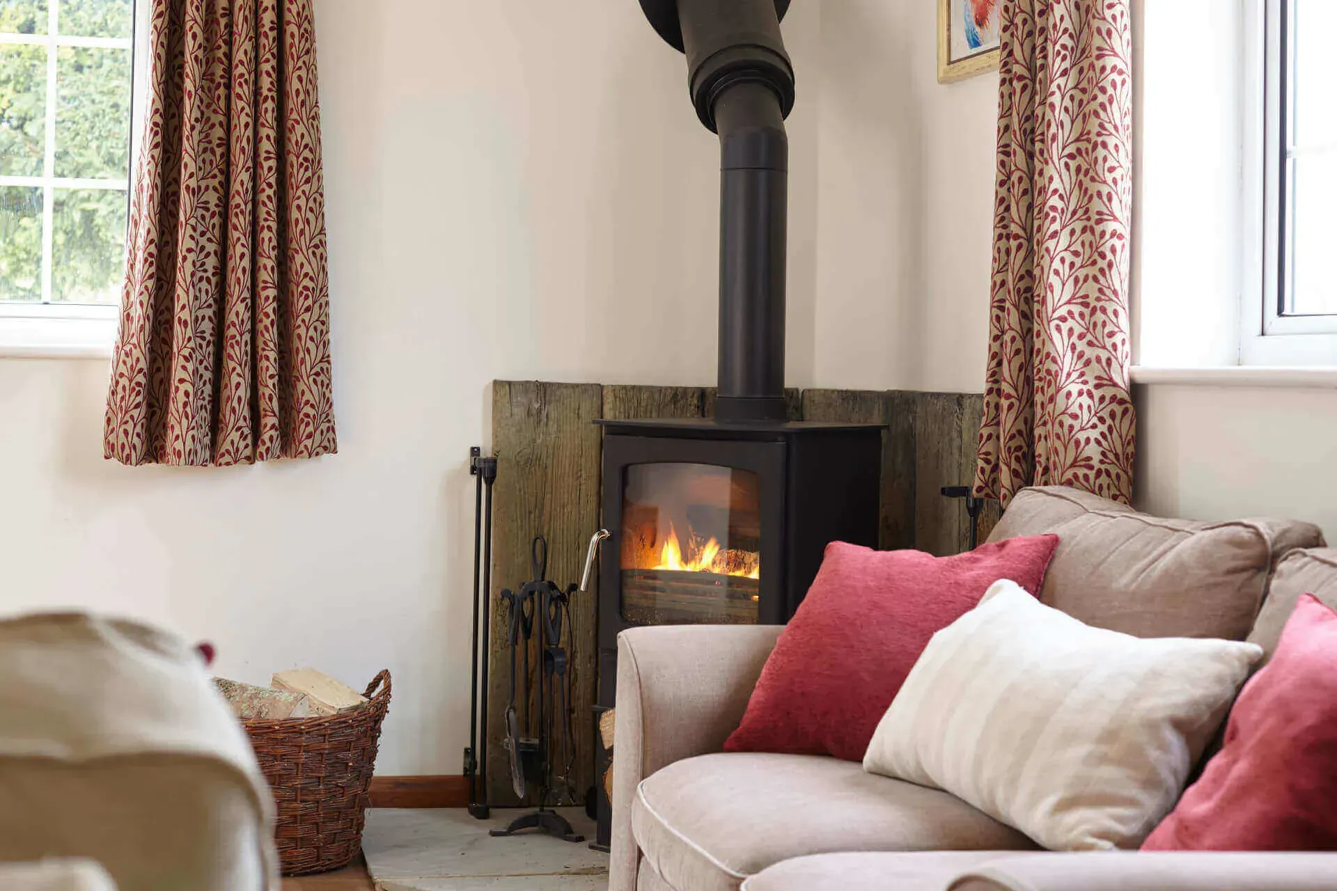 A wood-burning stove glows between two windows with patterned curtains, next to a sofa with red and white cushions.