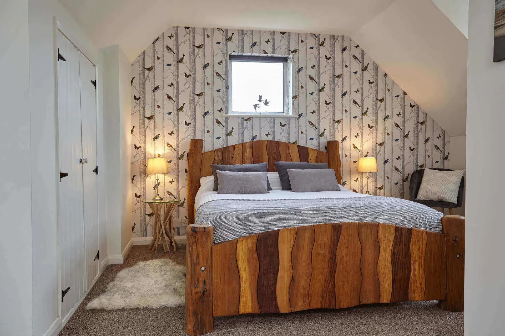 A bedroom with a rustic wood bed frame, patterned birch wallpaper, white closet doors, and two bedside lamps.