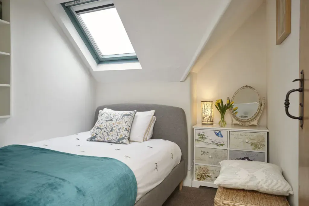 A cozy attic bedroom with a gray bed, teal throw, skylight, and a floral-patterned chest of drawers with a vanity mirror.