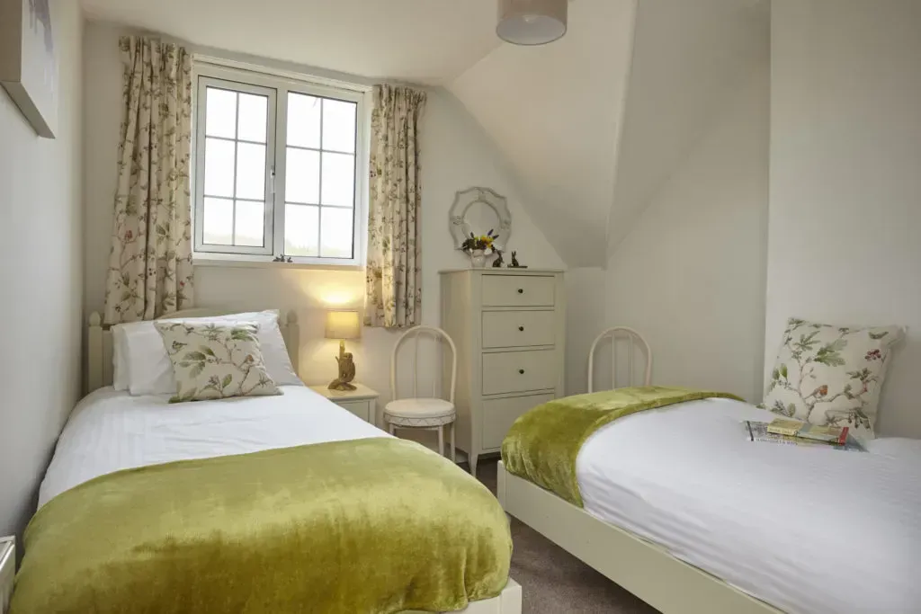 A bedroom with two single beds featuring white bedding, green throws, patterned pillows, a chest of drawers, and a window.