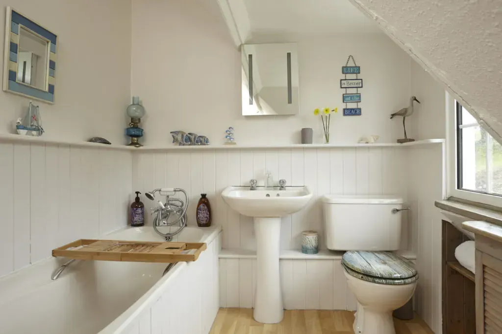 A bright bathroom with white beadboard walls, a bathtub, pedestal sink, toilet, and nautical-themed decor on a shelf.