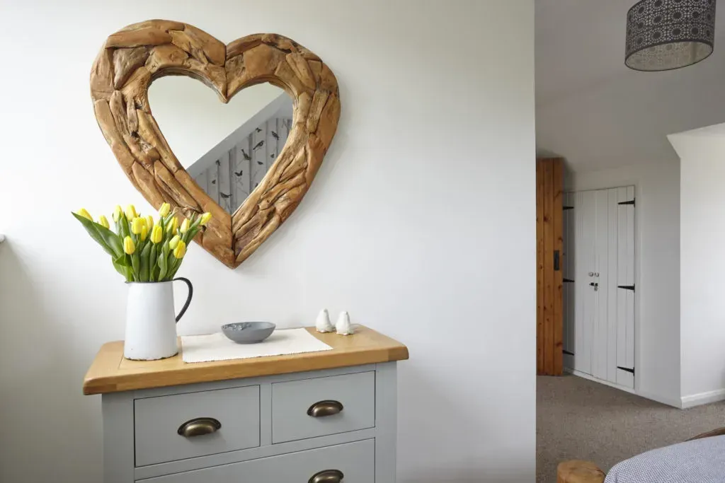 A grey chest of drawers with a heart-shaped wooden mirror above, topped with a white pitcher of yellow tulips.