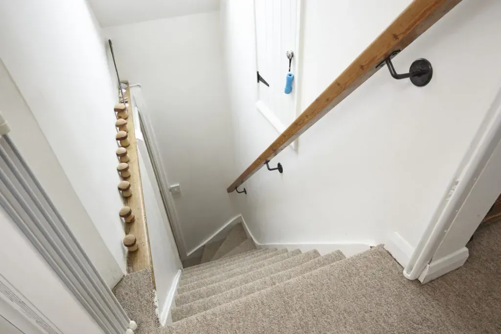 A downward view of carpeted stairs with a wooden handrail on the right and a wall-mounted wooden coat rack on the left.